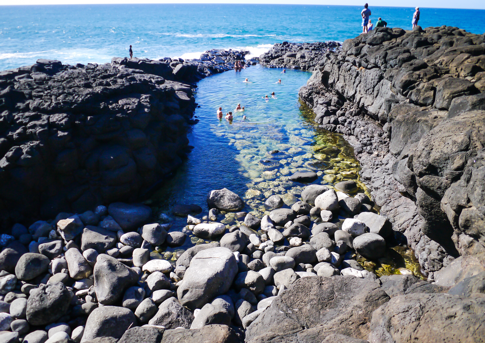Tourists stand on rocks and bathing in a water reservoir at Queen's Bath, its rocky coast is one of the best snorkeling spots in Kauai