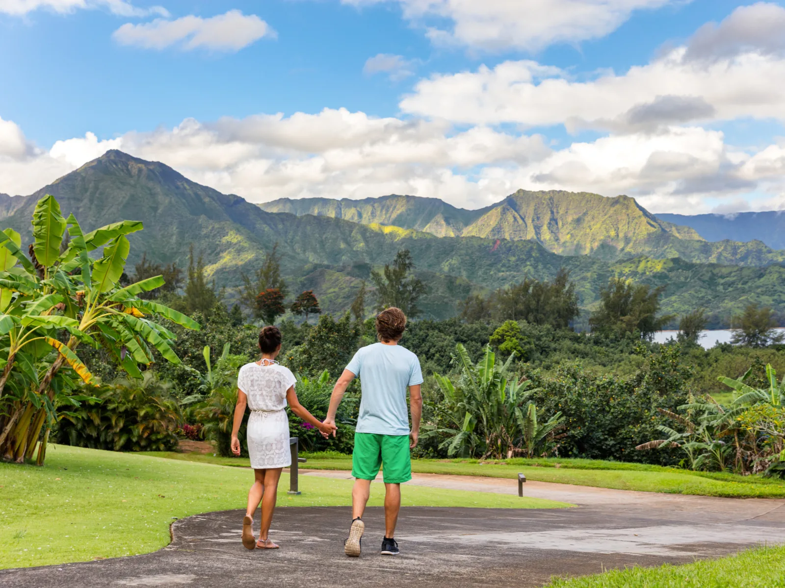 Attractive young couple in white walking along a path at one of the best hotels in Kauai with the volcanic mountains looming overhead in the distance