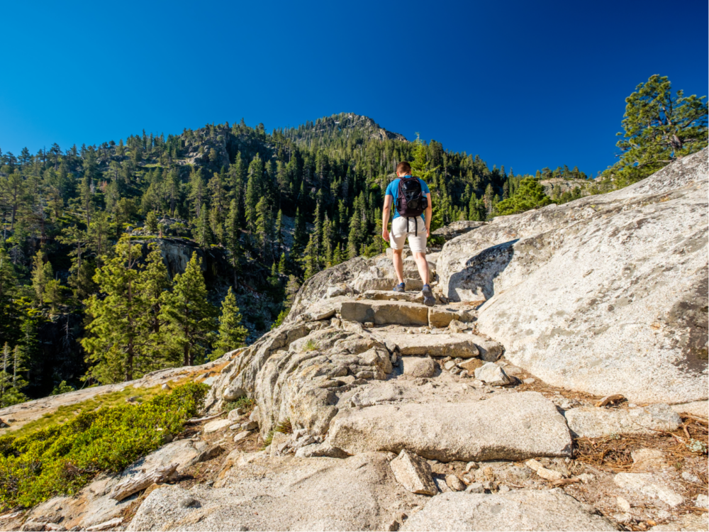 Man backpacking up the rocks on a mountain with pines all around during the best time to visit Lake Tahoe