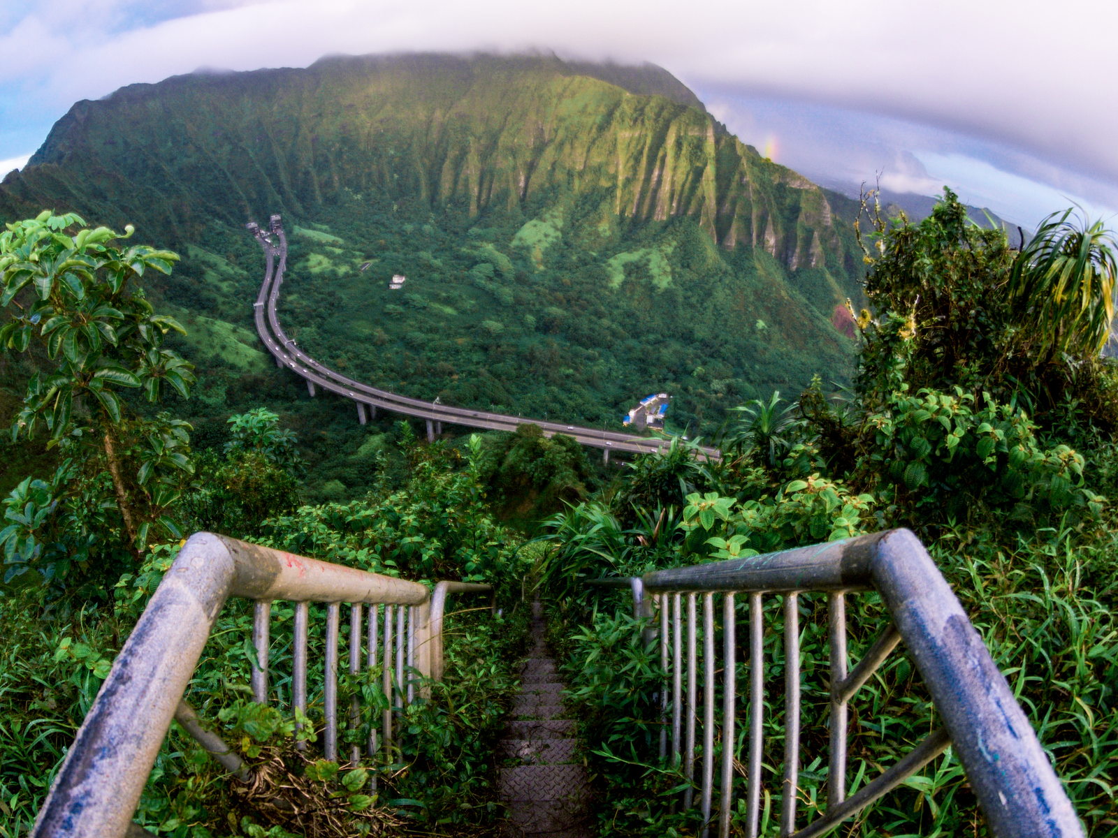 Photographed from the peak of Haiku Stairs, climbing here is one of the best things to do in Oahu, seen from below, a mountain tunnel connecting to a curved bridge over a thick forest