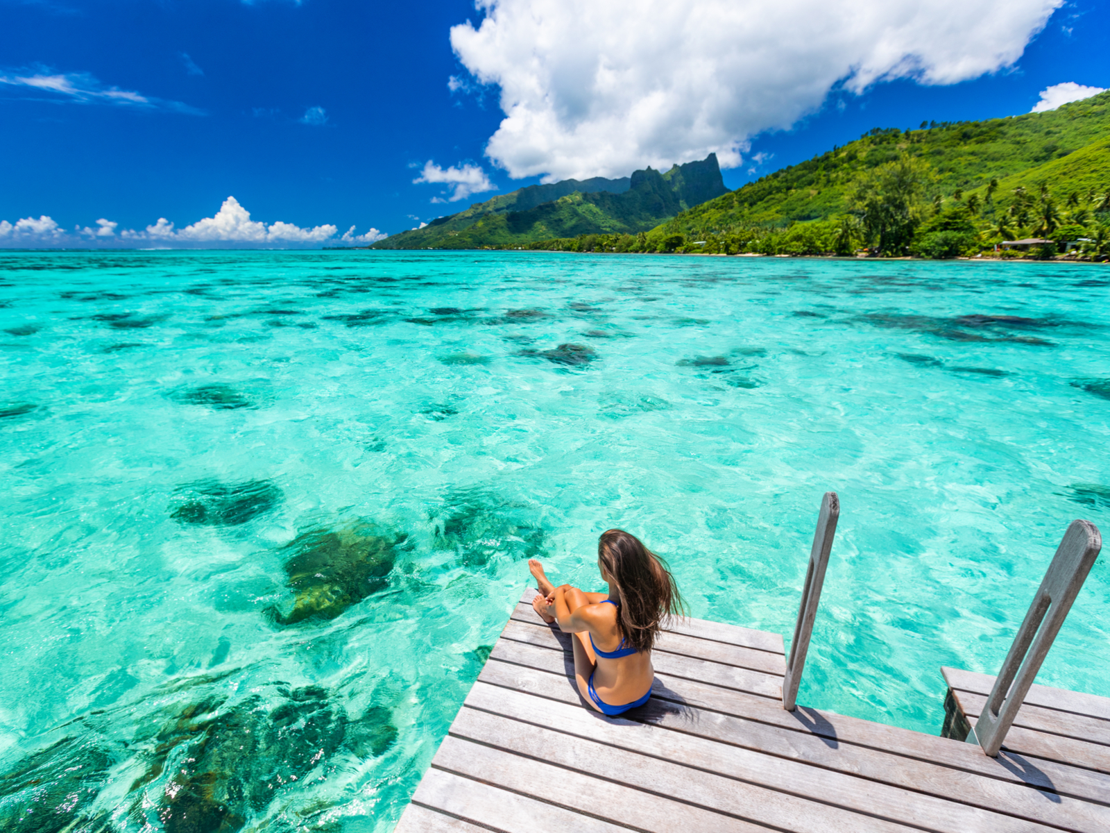 Woman in a blue bikini sitting on a dock over green water during the cheapest time to visit Tahiti