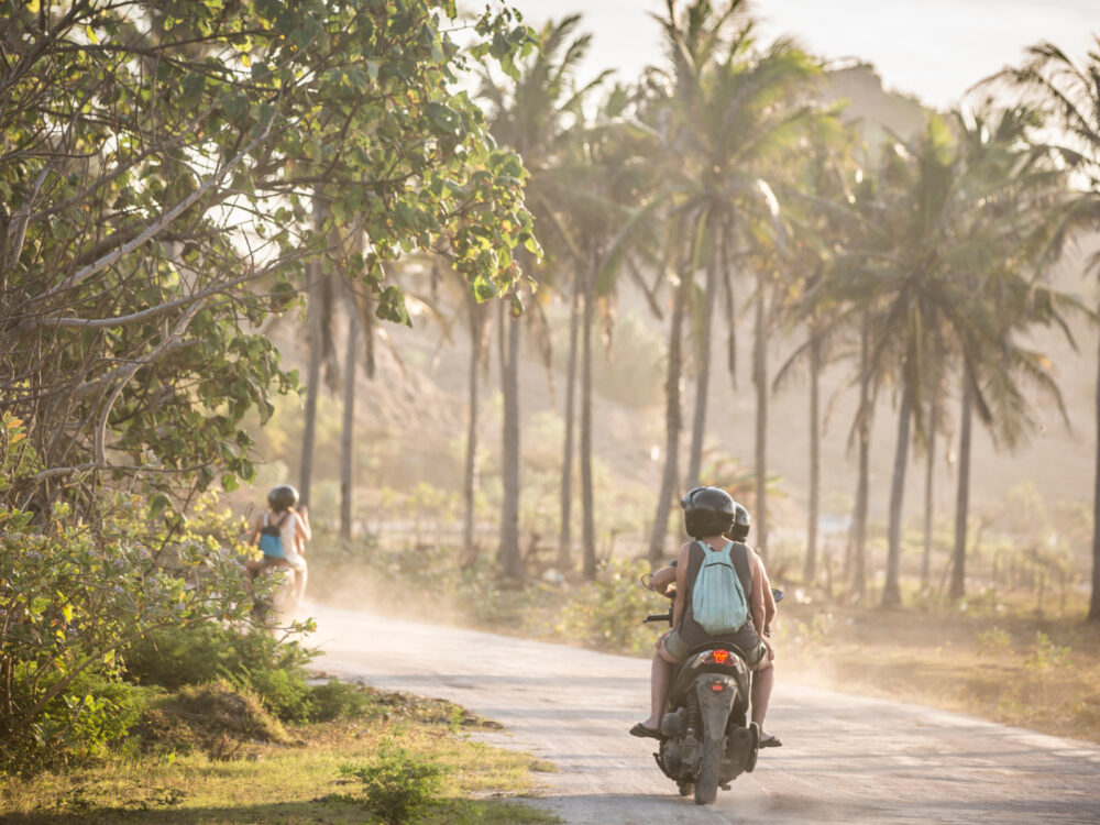 Two folks driving a scooter on a tropical dirt road for a piece titled Is Bali Safe