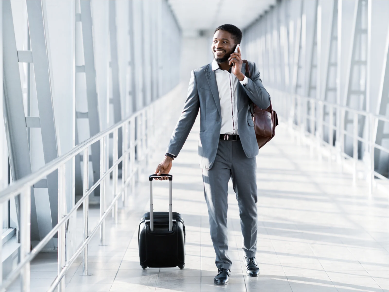 Front view of a man in a suit talking on a phone, toting a rolling suitcase, and wearing one of the best travel business backpacks