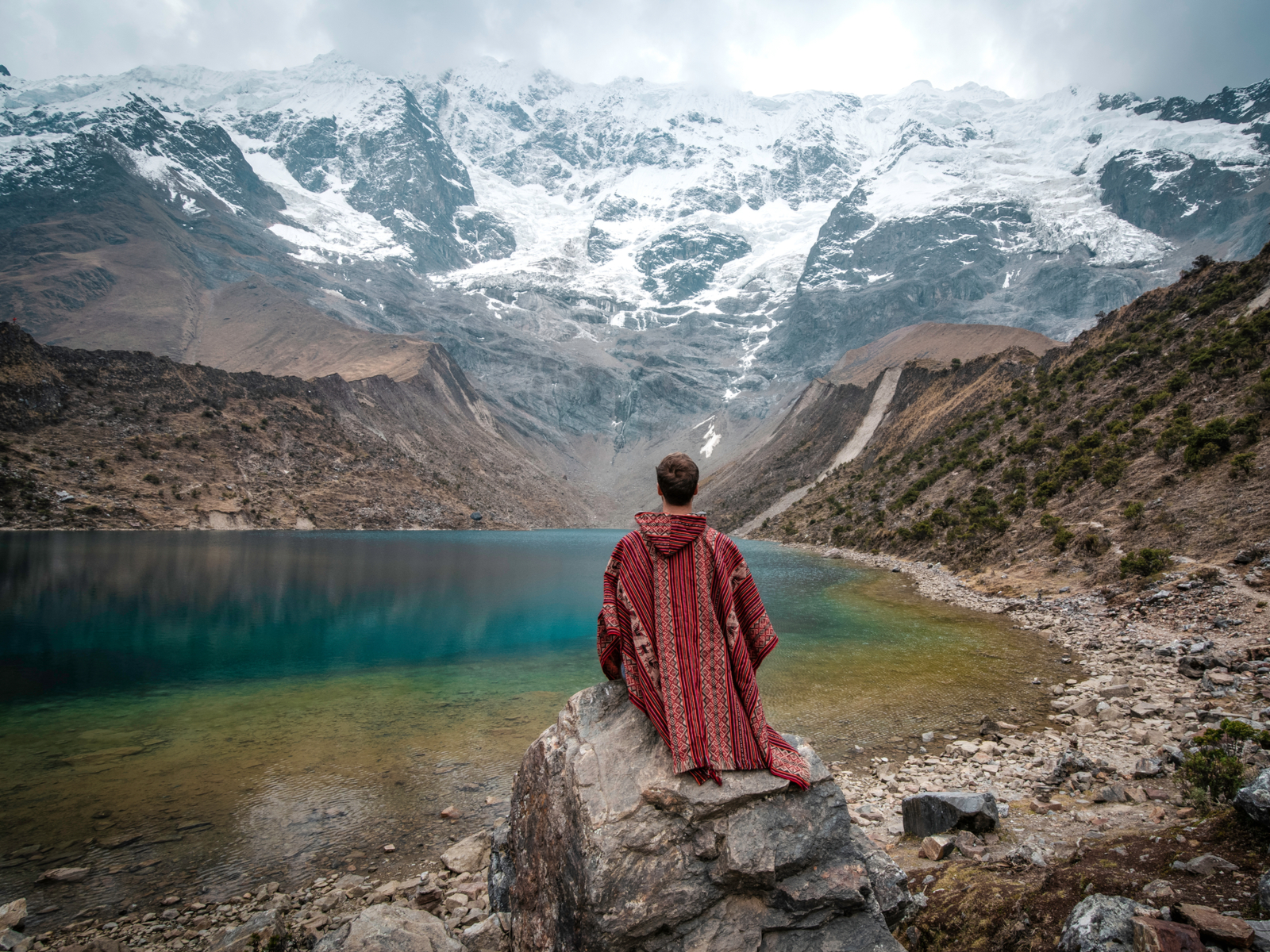 Young man in a poncho sitting on the banks of Laguna Humantay for a post titled Is Peru Safe to Visit