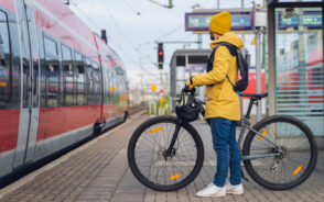 Guy in a yellow jacket with a pack on his back holds a bike by a train station for a piece title The Best Commuter Backpacks
