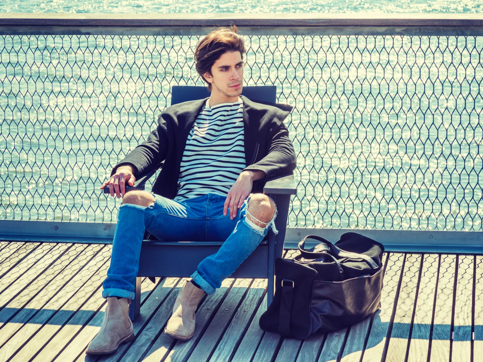 Stylish young man with ripped skinny jeans and a black men's leather weekender bag looking seriously to his left while sitting on a dock