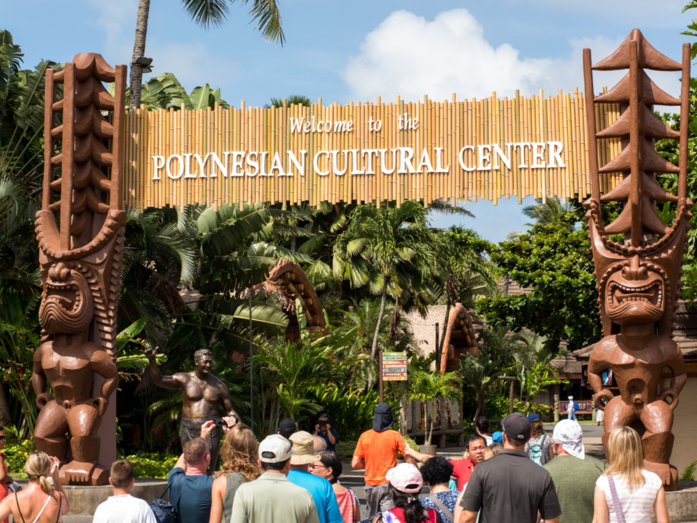 Tourists heading inside the Polynesian Cultural Center, one of the best things to do in Oahu, with two large tiki statues supporting the welcome arch