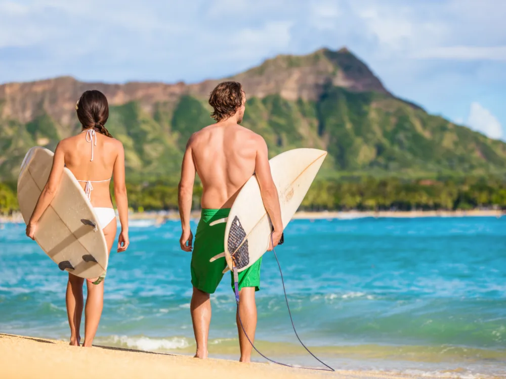 A couple in their swimwear carrying surfboards as they walk towards the wavy Waikiki Beach, among the best things to do in Oahu, with the famous Diamond Head Mountain blurred in the background