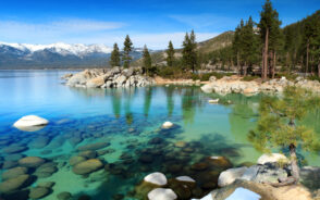 Cover photo for a piece titled Where to Stay in Lake Tahoe featuring a clear teal lake with rocks at the bottom with snowcapped mountains in the background
