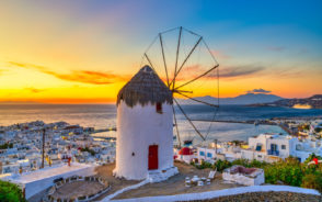 Featured image for a guide on where to stay in Mykonos featuring a windmill overlooking the ocean and town at sunset