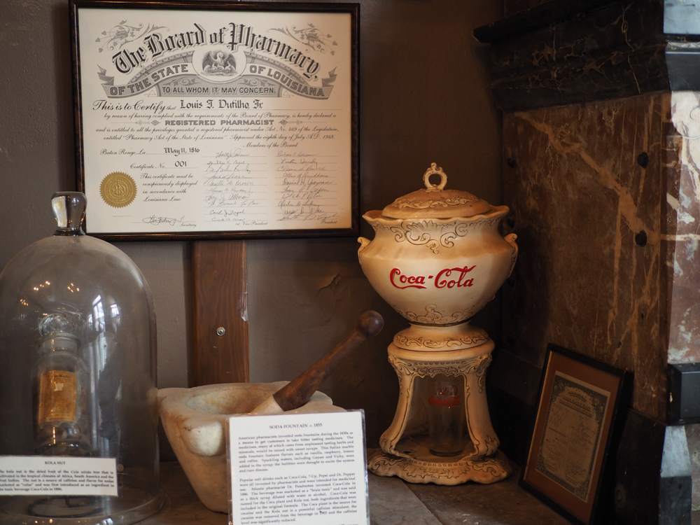 Old items displayed in New Orleans Pharmacy Museum shelf where one item can be seen as an old ceramic pot that has the brand of Coca-Cola on it, visiting the museum is one of the best things to do in town.