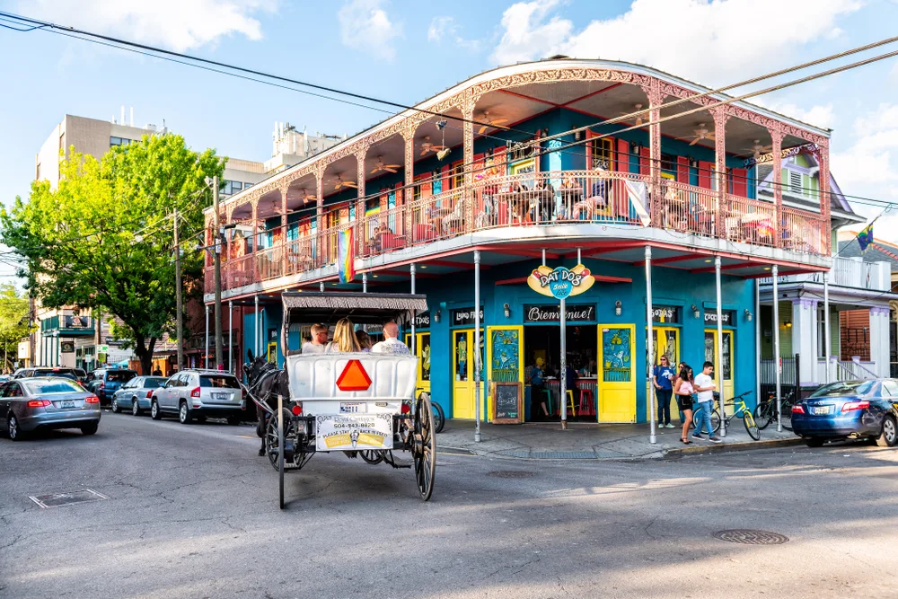 A two-story old building at the corner of a block where a horse carriage can be seen with passengers and cars parked on the side of the street, one of the historic destination on our list of things to do in New Orleans.