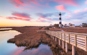 Featured image for a piece titled The Best Time to Visit the Outer Banks with a lighthouse in the middle of a marsh and a wooden walkway leading up to it