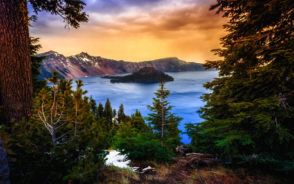 View of a lake and a bunch of trees as seen from the top of the mountain during the best time to visit Crater Lake with the sun setting behind the mountain