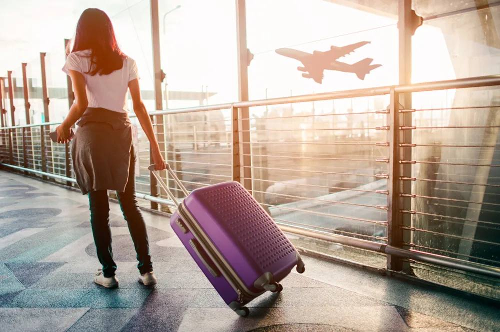 Woman pulls her purple luggage behind her walking through an airport with an airplane seen taking off through the window