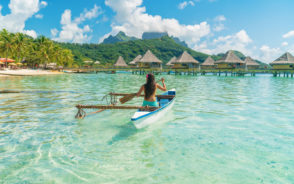 Woman in Bora Bora pictured paddling her wooden boat on the crystal-clear water by the bungalows during the best time to visit French Polynesia