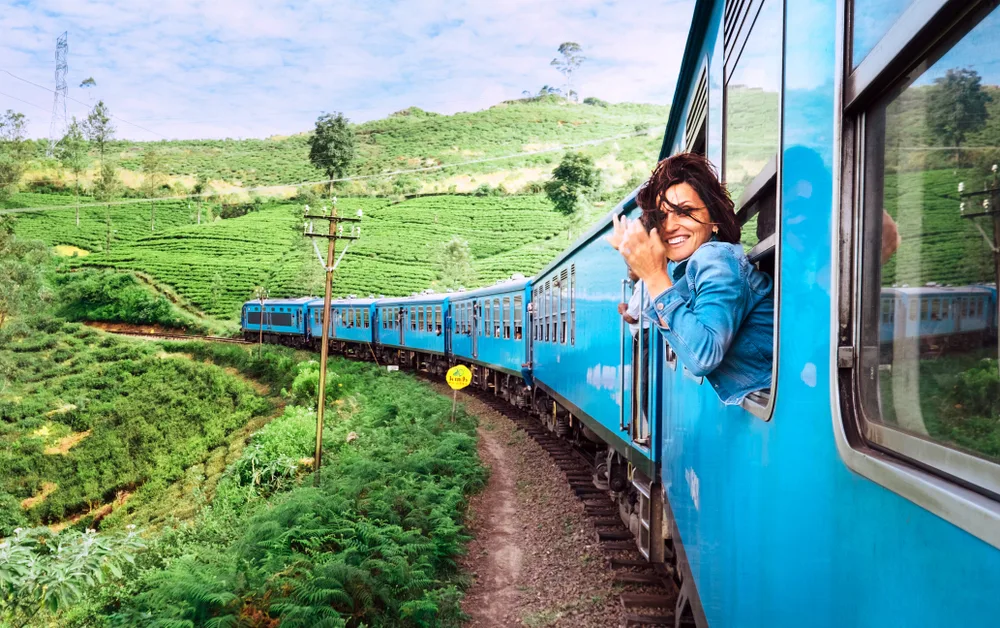 Smiling woman looking out of the window of a blue train wearing a blue jacket while it goes through rice fields in Sri Lanka