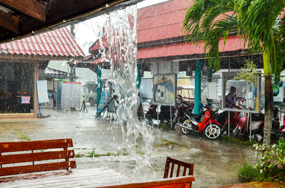 Heavy rainfall cascading off a building during the overall worst time to visit Phuket with a scooter outside in the rain and muddy walkways outside