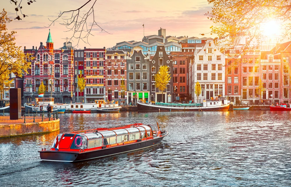Boat in Amsterdam cruising on the river during dusk with the sun setting over the historical and colorful buildings