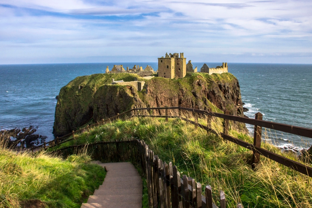 Dunnottar Castle pictured on a coastline with a few clouds in the sky and waves lapping the shore during the overall best time to visit the United Kingdom
