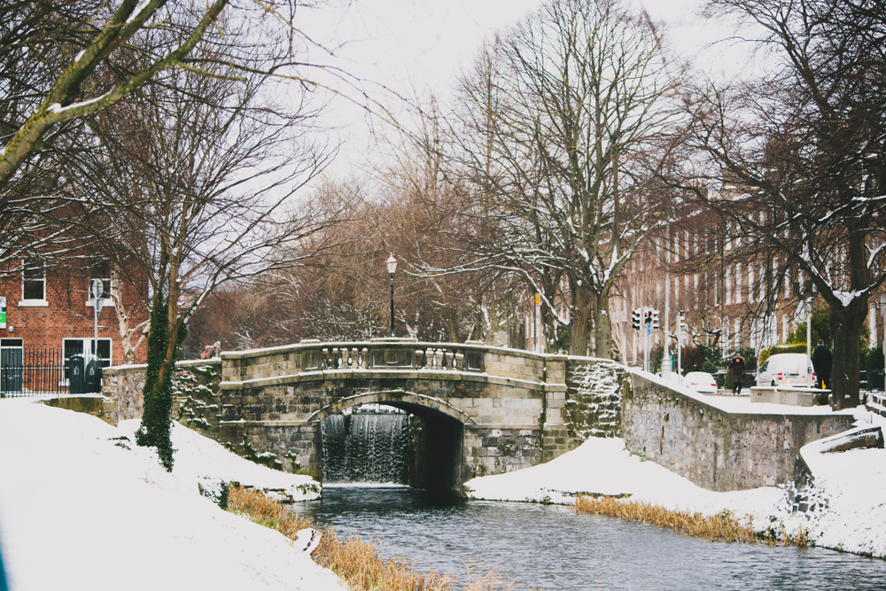 A stone bridge in Dublin pictured during the cheapest time to visit with a gloomy sky and snow on the ground with trees, sans leaves