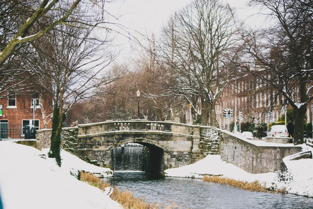 A stone bridge in Dublin pictured during the cheapest time to visit with a gloomy sky and snow on the ground with trees, sans leaves