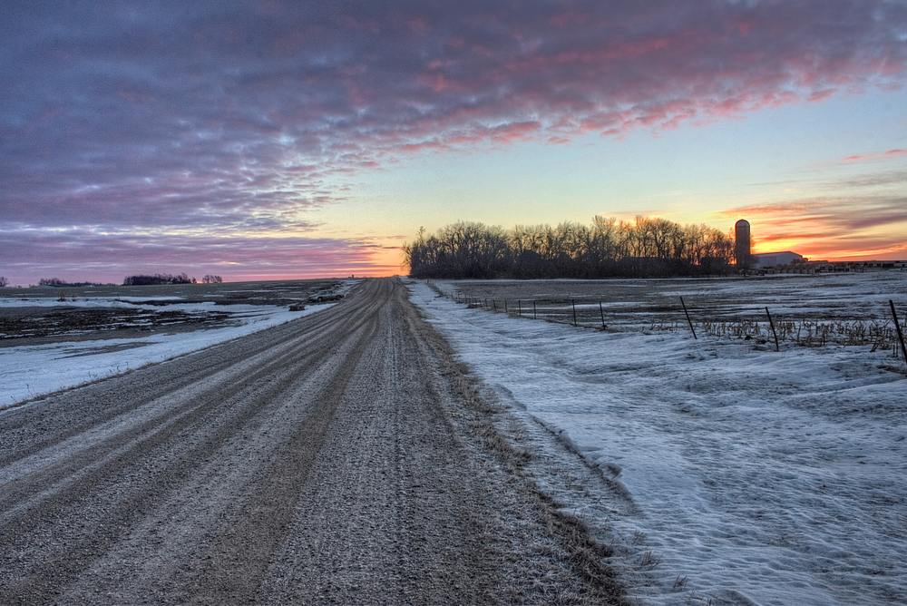 Image of a dirt road with snow on either side and a gorgeous sunset over the trees and behind a farmhouse pictured during the winter, the worst time to visit South Dakota