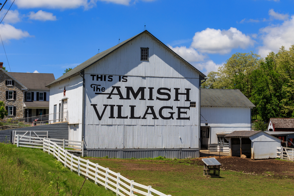 White barn that says This Is The Amish Village on a blue sky day