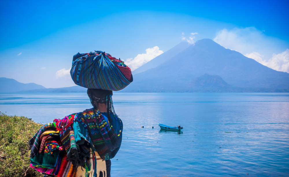 Woman depicted carrying a basket on her head during the cheapest time to visit Guatemala and looking out over the ocean with a volcano in the background