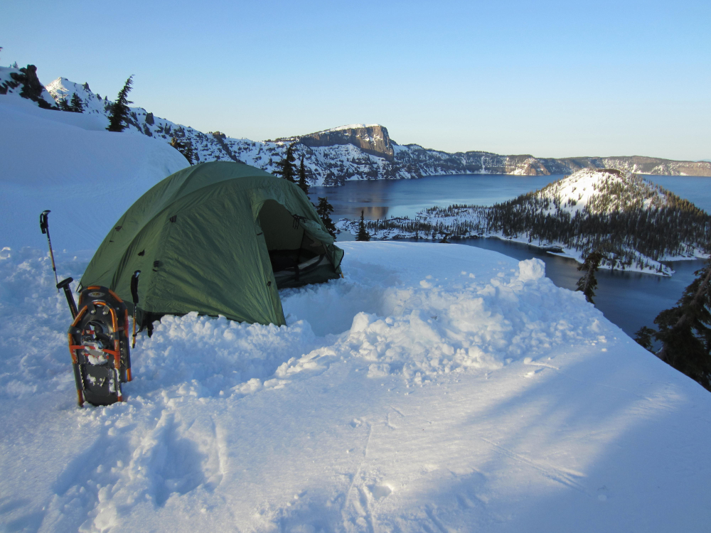 Person camping on a hill in Crater Lake during the winter, the cheapest time to visit, with snowshoes and poles stuck in the ground next to their tent