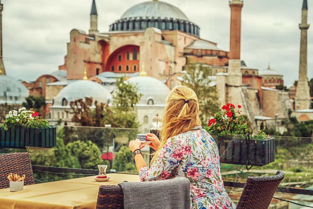 Woman drinking Turkish coffee while wearing a floral robe and sitting on a balcony overlooking the mosque during the best time to visit Istanbul, the summer