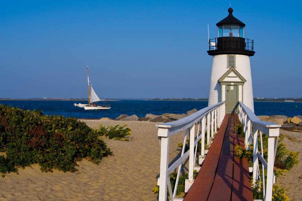 Neat view of the ocean at the base of a walkway to a lighthouse with a sailboat in the background for a piece on the best time to visit Cape Cod