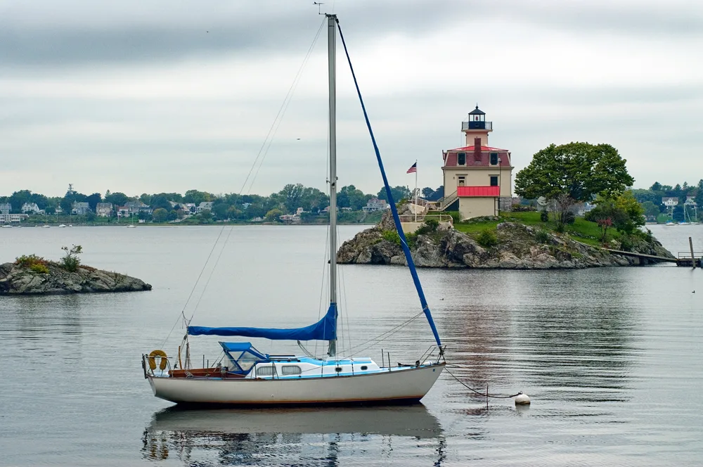 Pomham Rock Lighthouse with blue sailboat in the water on a cloudy day for a frequently asked questions section on the best time to visit Rhode Island