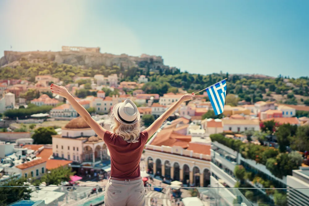 Woman standing on a hill and holding up a Greek flag in her right hand while overlooking the valley