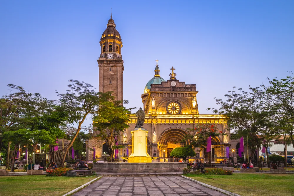 Beautiful view of the cathedral at Manila pictured during the best time to visit the Philippines with the front lit up by yellow light and a rustic brick path leading to a fountain