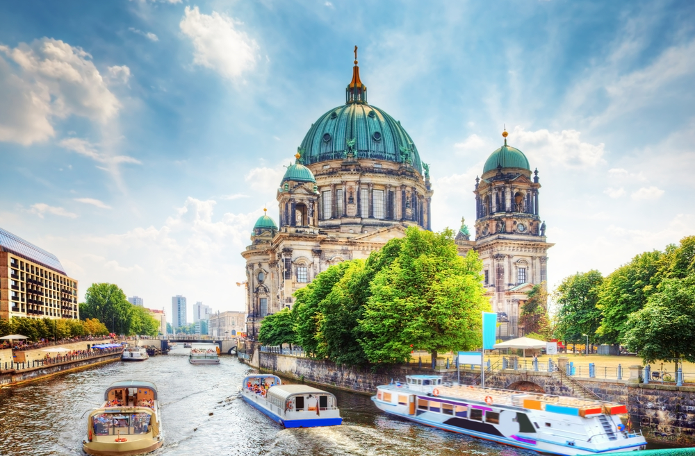 Berlin Cathedral during the best time to visit Berlin with green trees, sunny, cloudy skies, and boats in the water