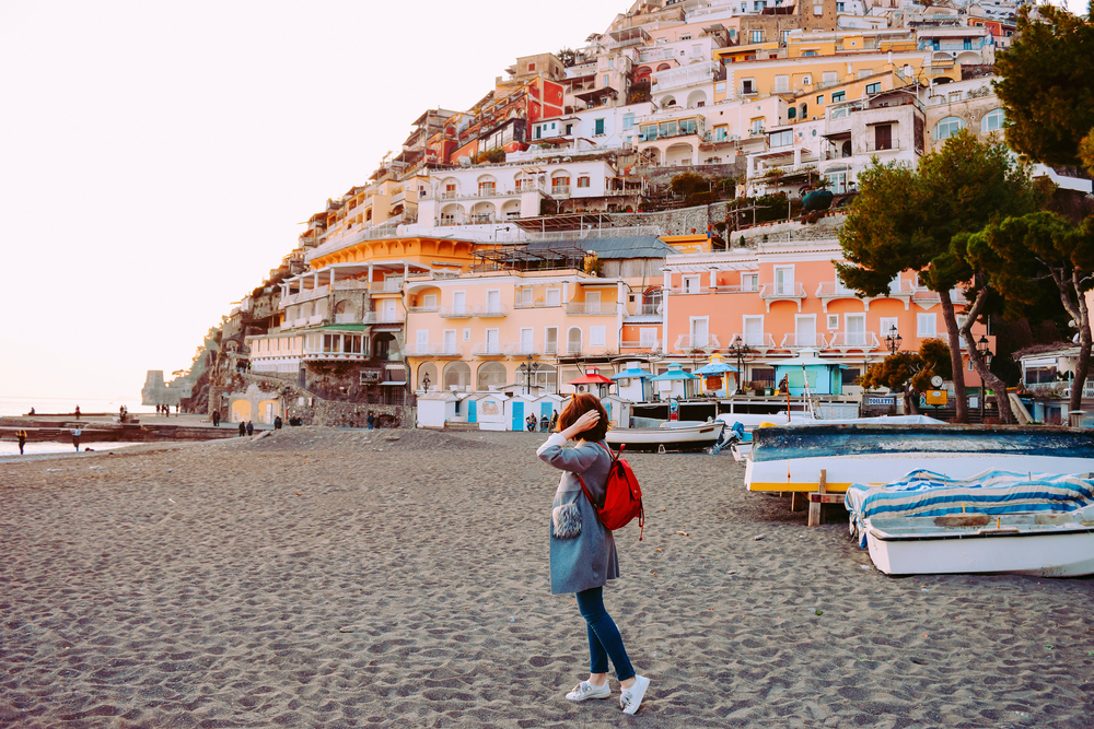 Woman in a backpack, pants and a jacket standing on a beach in Positano during the off-season, the cheapest time to visit the Amalfi Coast