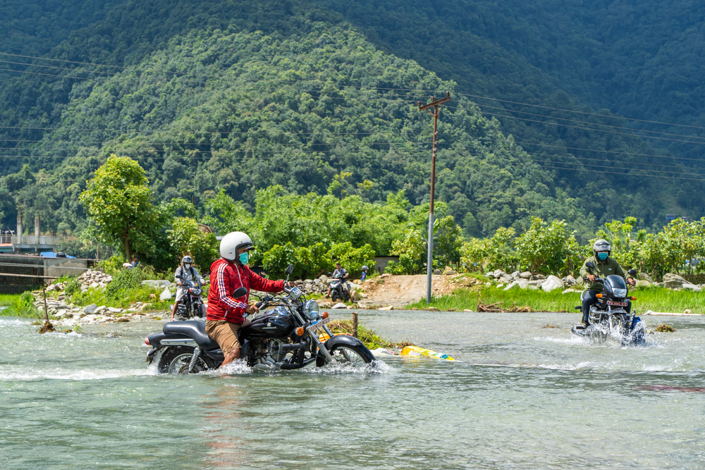 Two guys pictured riding bikes through water with a giant mountain in the background during the monsoon season, the cheapest time to visit Nepal