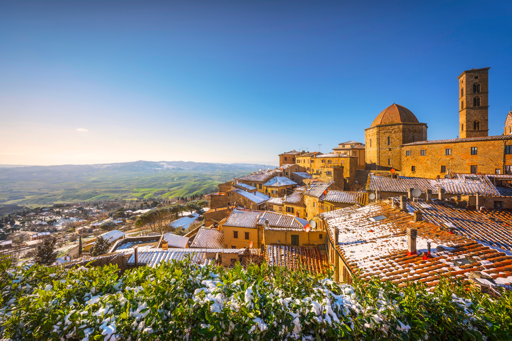Volterra pictured during the winter, the worst time to visit Tuscany, with snow on the ground and bushes and roof while the town overlooks a valley which is covered by a deep blue sky