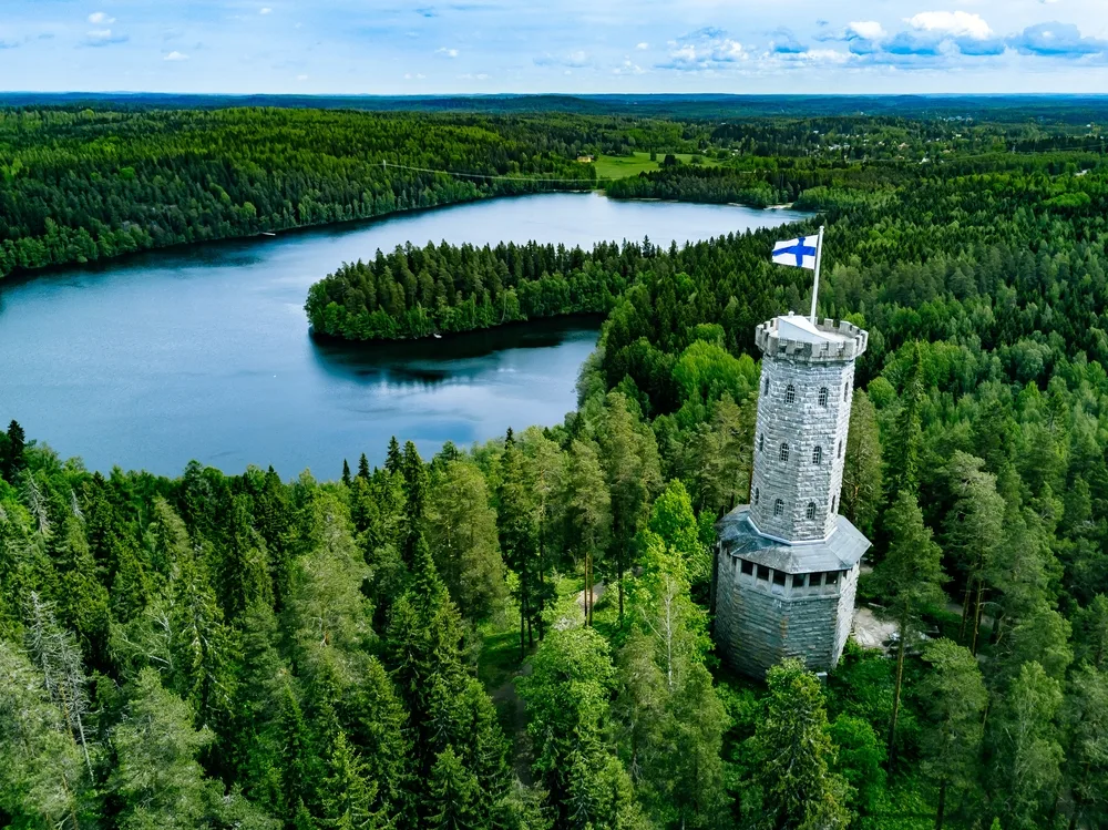 Aerial image of a stone observation tower protruding up from a green forest and flying a Finnish flag