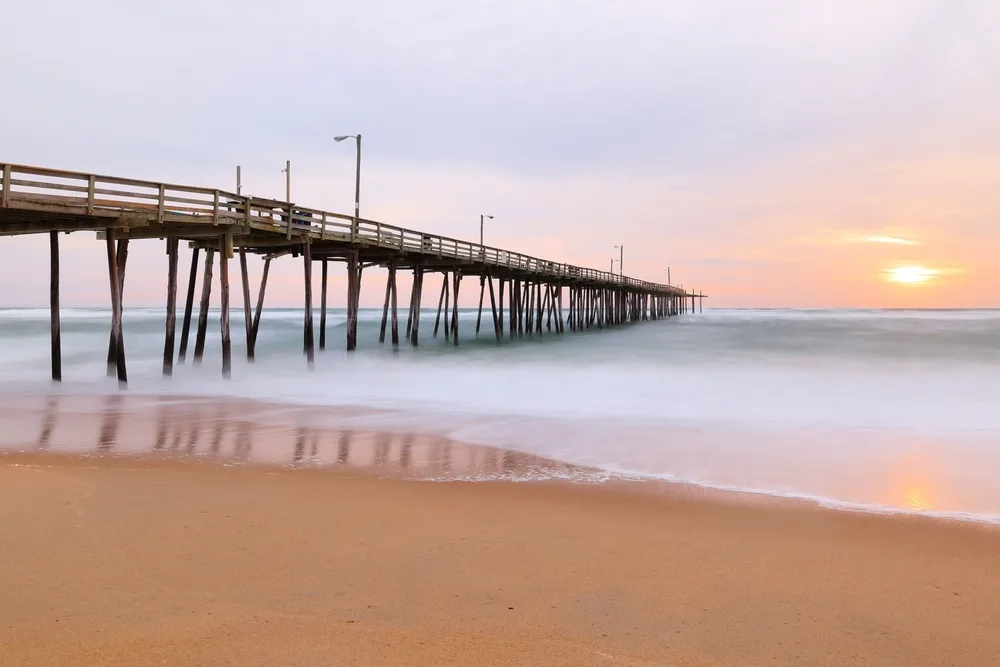 Waves lap up against the footings of a pier next to a brown sand beach in the Outer Banks