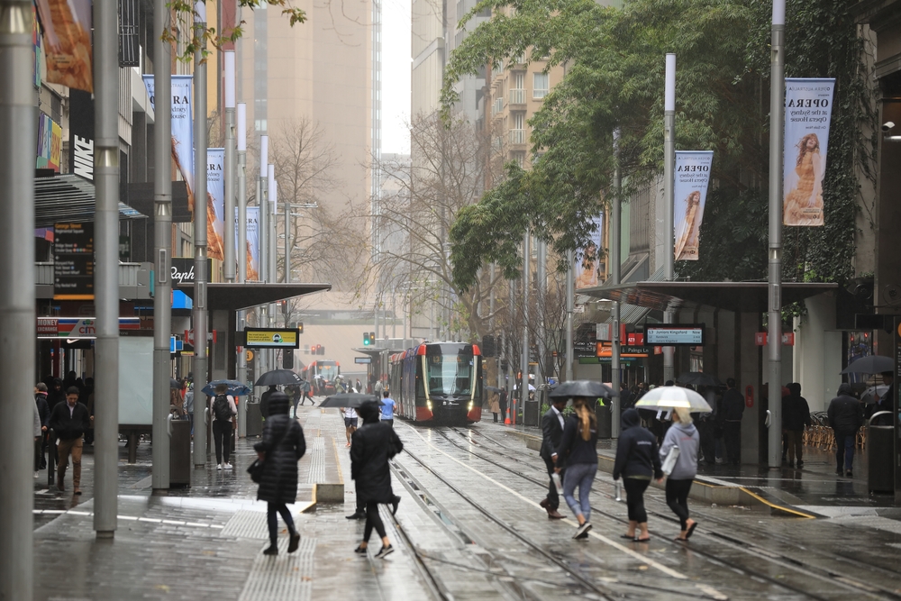 People walking through downtown Sydney with rain falling on them and a tram in the middle of the street making its way toward the camera during the rainy season, the cheapest time to visit Sydney