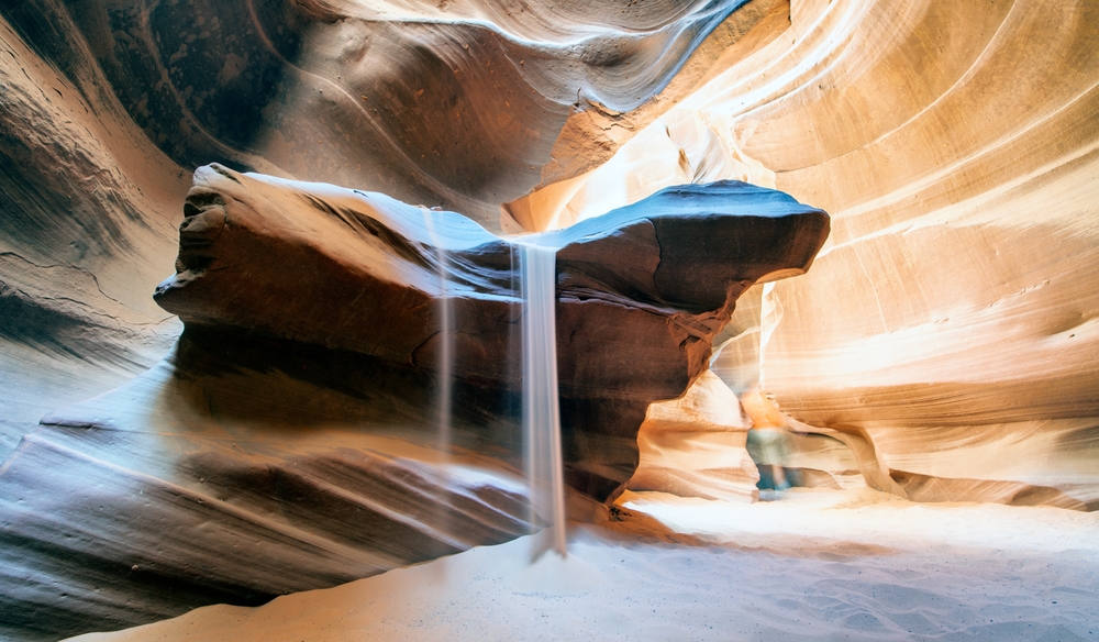 Sand running down a giant rocky ledge with the sun poking its head in during the best time to go to Antelope Canyon
