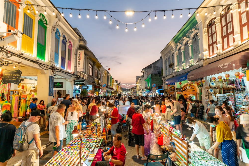 Photo of people swarming a market in the spring, the overall best time to visit Phuket, with yellow and brown buildings on either side of a crowded marketplace with tents and lights, as seen at dusk