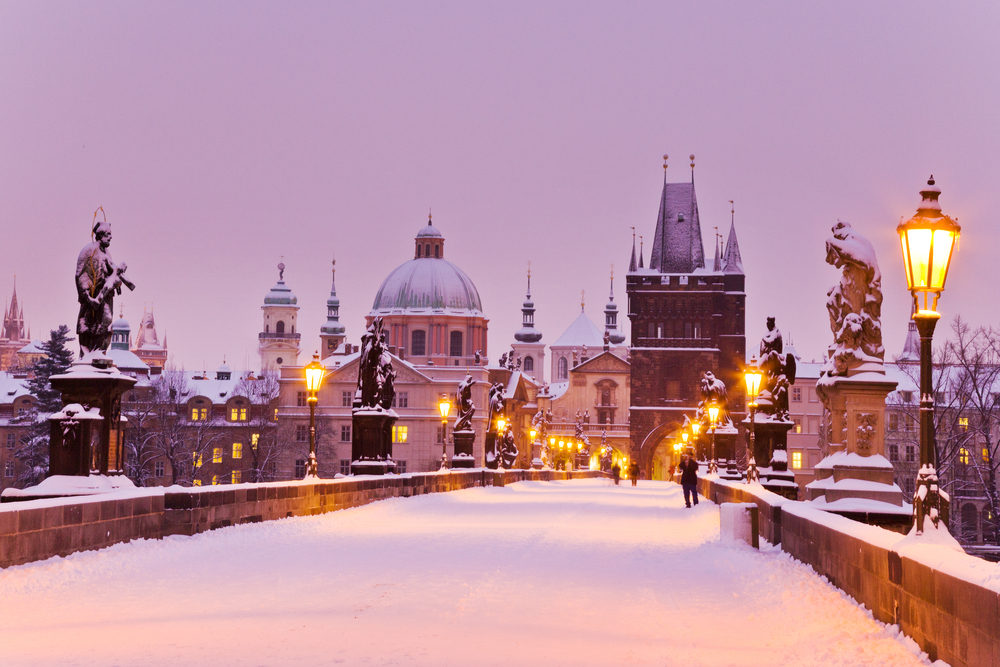 Winter in Prague, also considered the cheapest time to visit, pictured with a snowy bridge leading to the town