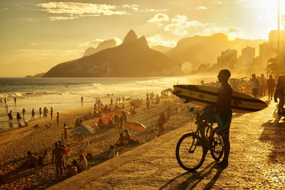 Guy with a surfboard pictured on a beach at dusk and putting his foot on a bike for a piece on the best time to visit Rio de Janeiro