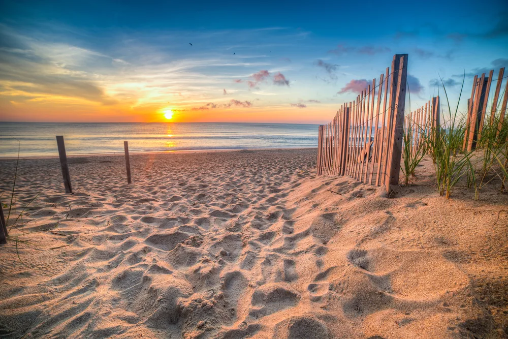 A gorgeous and serene sand dune viewed from a walkway with wooden dune barriers overlooking the ocean while the sun sets during the best time to go to the Outer Banks