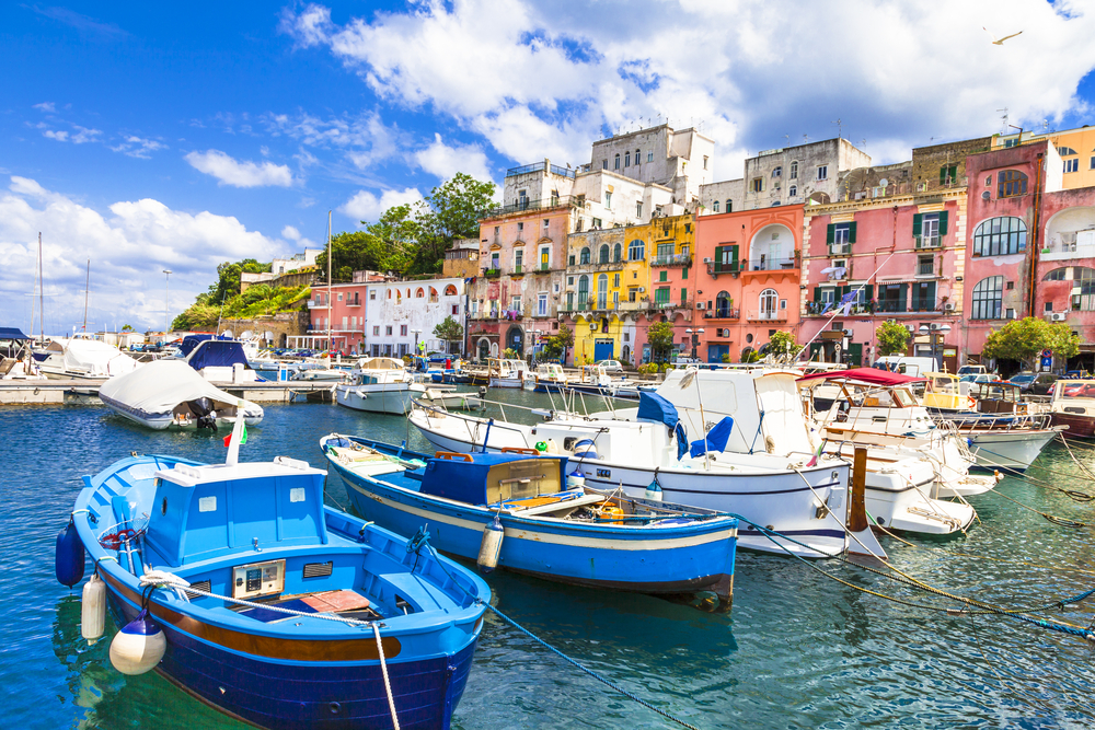 Gorgeous and colorful boats in the bay of Procida with colorful buildings lining the cliffs above for a piece on the best time to visit the Amalfi Coast
