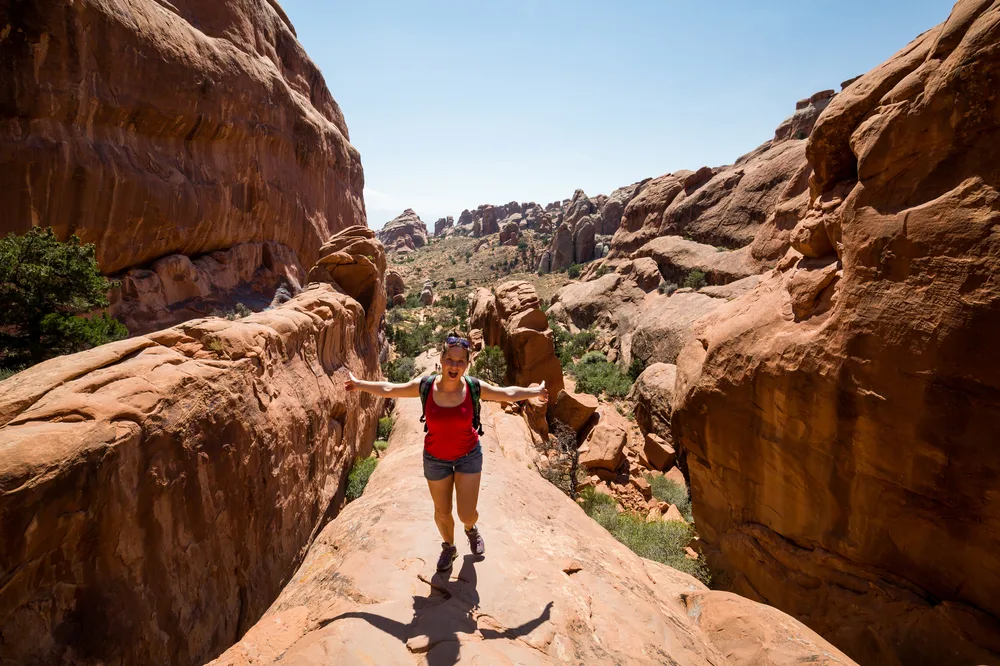Woman in hiking shorts wearing a backpack pictured holding her arms out toward either side of the canyon during the best time to visit Arches National Park