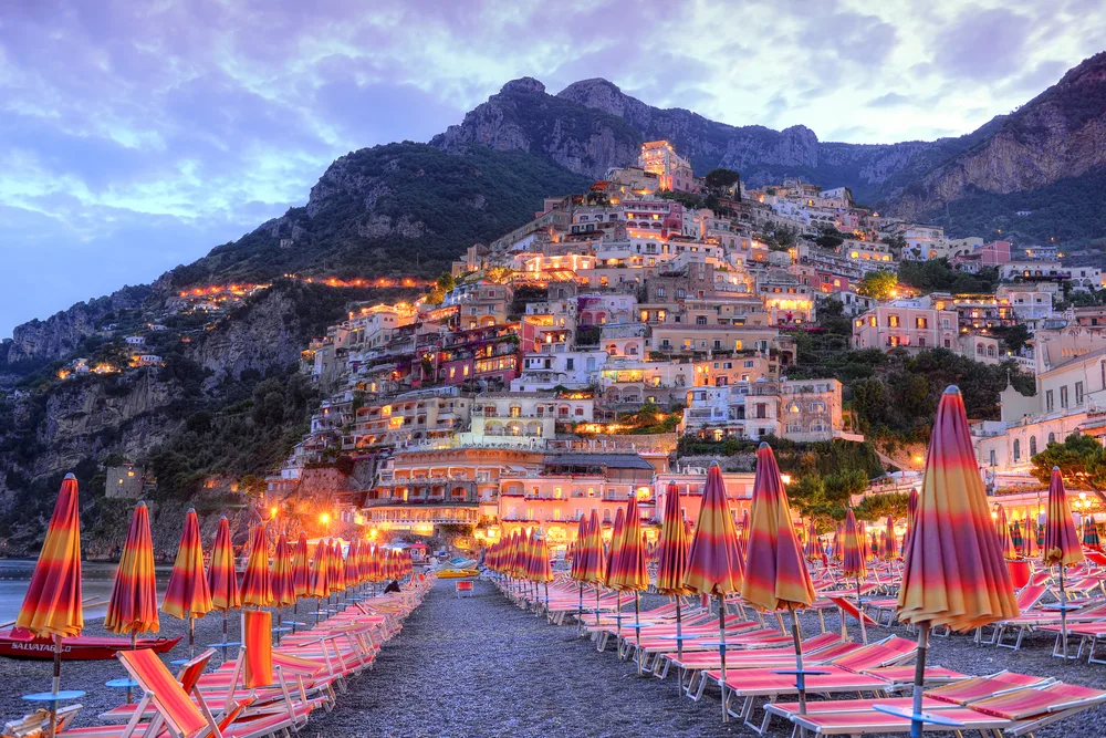 Night view of Positano pictured during the best time to visit the Amalfi Coast with lights and pink and red umbrellas on the sand